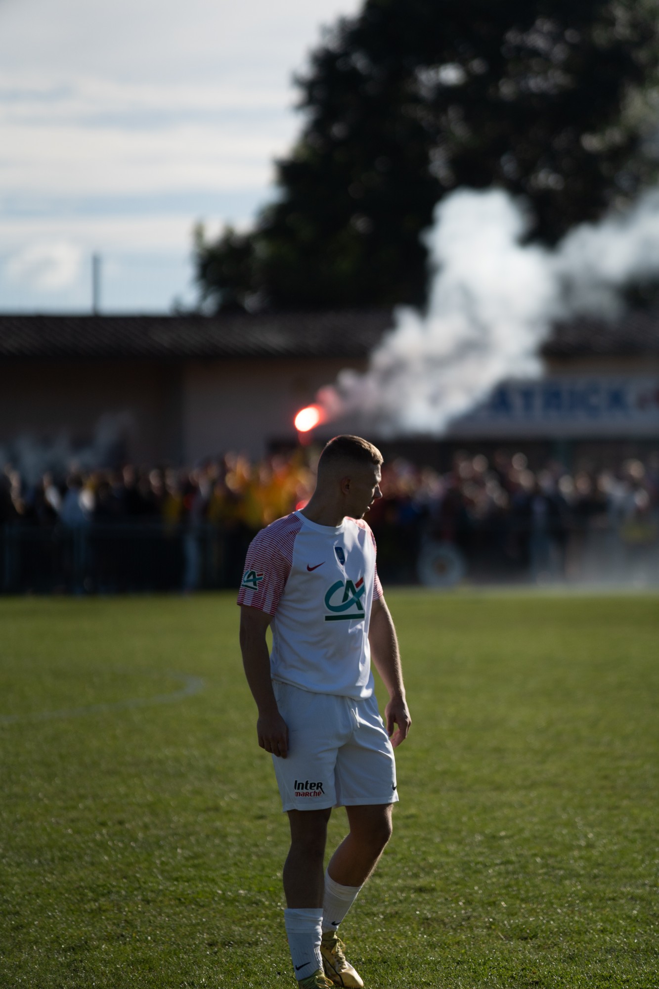 Footballeur en maillot blanc sur la pelouse, fumigène et supporters en arrière-plan